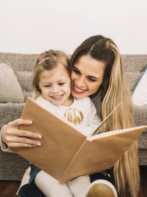 smiling-mother-daughter-reading-book-near-couch smiling-mother-daughter-reading-book-near-couch